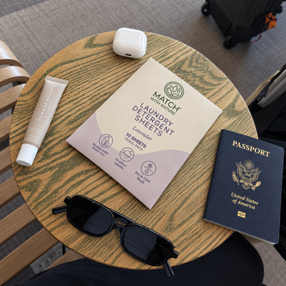Lavender Travel Pack on a café table at the airport, beside a passport, sunglasses, an earbuds case and lip balm